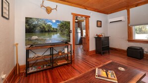 A room with a wooden ceiling, a large painting of a river, a wooden coffee table with a book, and a kitchen area visible through an open doorway. - Fishing, Kayaking, Pet-friendly on the Smith River (Bassett)