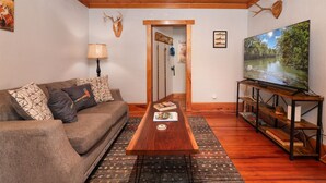 A cozy living room with a wooden ceiling, a brown sofa with patterned cushions, a coffee table with books, a flat-screen TV on a wooden stand, and a door leading to another room.