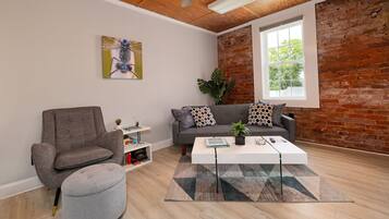 En moderne stue med en grå lænestol, et hvidt sofabord og en grå skammel. Der er en grå sofa med mønstrede puder, en stor plante og et vindue med udsigt til træer.