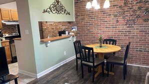 Un'area pranzo con un tavolo rotondo in legno e sedie, una cucina sullo sfondo e un muro in mattoni con decorazioni in metallo.