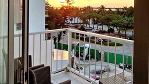 Un balcón con mobiliario de mimbre, vistas a un atardecer y una valla verde al fondo.