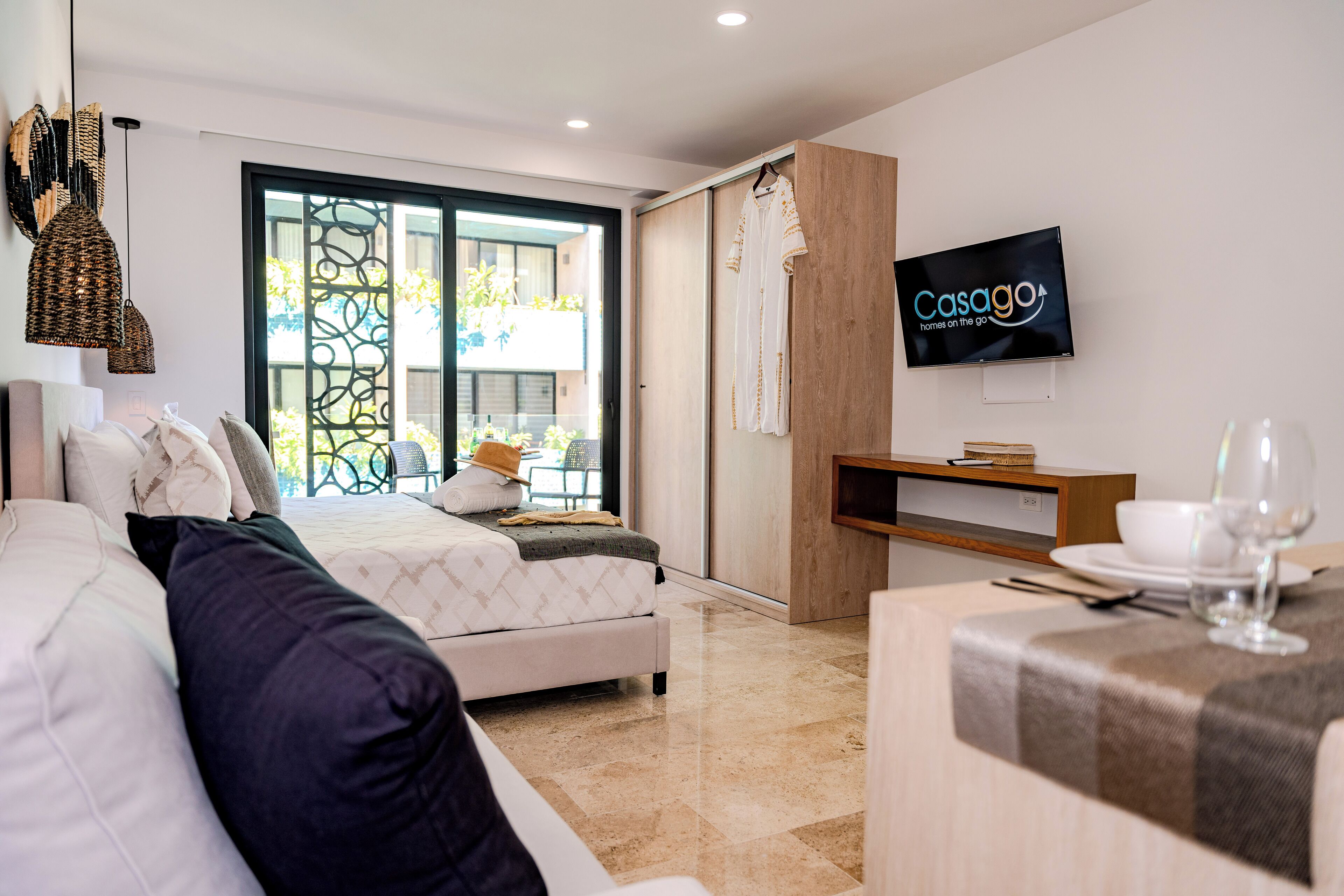 A modern hotel room with a bed, a TV, a dining area, and a balcony with a view.