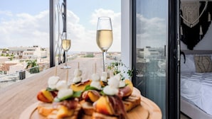 Un balcón con una mesa de madera dispuesta con aperitivos y una copa de vino blanco, con vistas a un paisaje urbano.