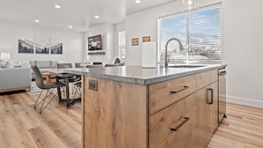 Una cocina moderna con una isla central, un comedor con un sofá y sillas grises, y un amplio ventanal con persianas.