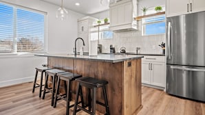 Una cocina moderna con una isla central, taburetes de bar, electrodomésticos de acero inoxidable y un ventanal con persianas.