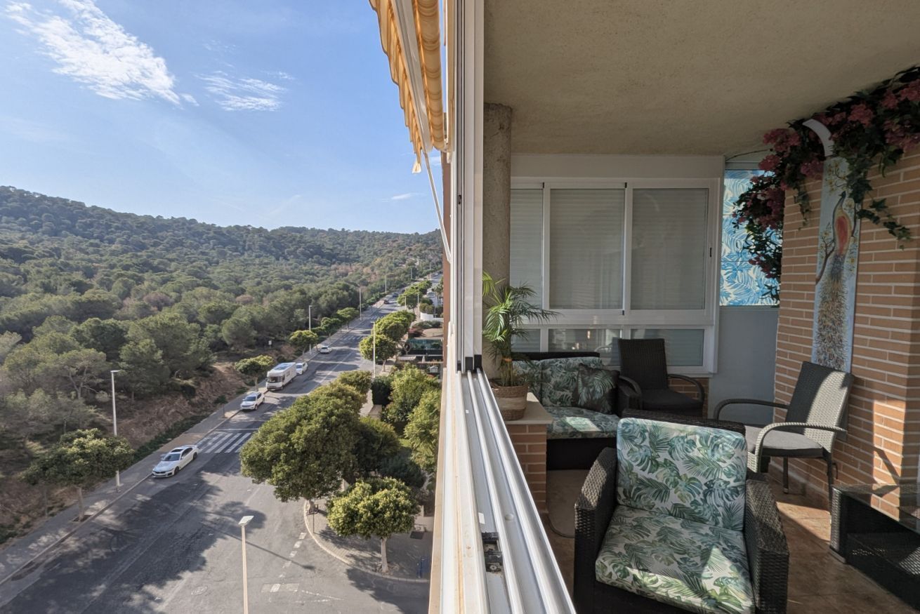 Un balcon donnant sur une route et des collines, des meubles en osier et une fenêtre agrémentée d’un rideau décoratif.