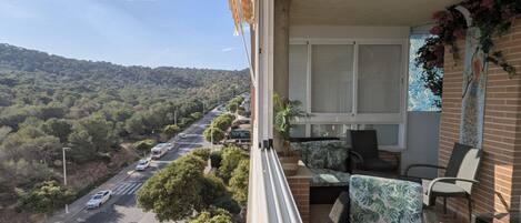 Un balcon donnant sur une route et des collines, des meubles en osier et une fenêtre agrémentée d’un rideau décoratif.