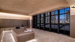 Une chambre d’hôtel moderne dotée d’une grande fenêtre donnant sur la ville, d’un aménagement spacieux et d’un coin salon central avec une petite table et un porte-serviettes.