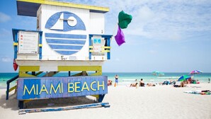 Une tour de surveillance avec l’inscription « MIAMI BEACH » à la base, un drapeau d’avertissement sur la plage et un parasol en arrière-plan.