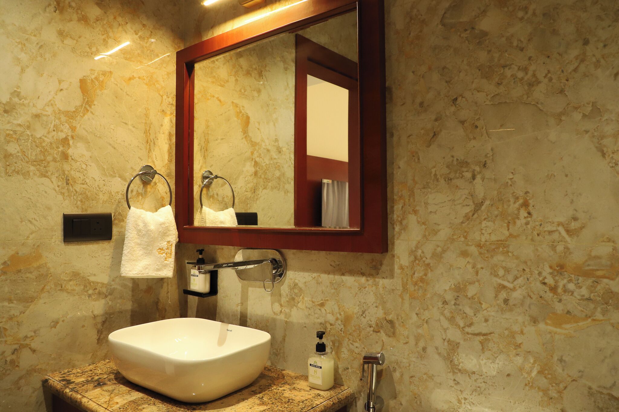A bathroom with a marble wall, a white sink, a towel rack, a mirror, and a soap dispenser.