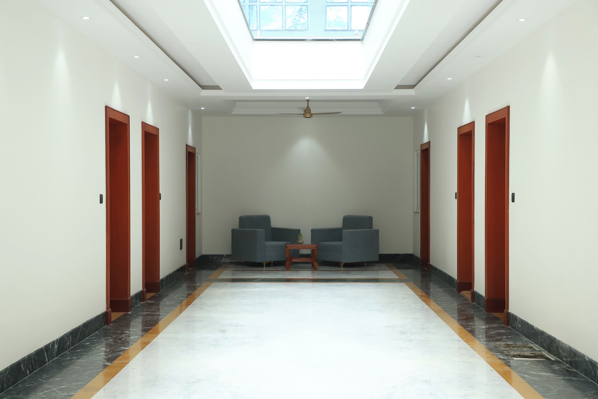 Um corredor com claraboia, duas poltronas, uma mesa pequena e portas de madeira.