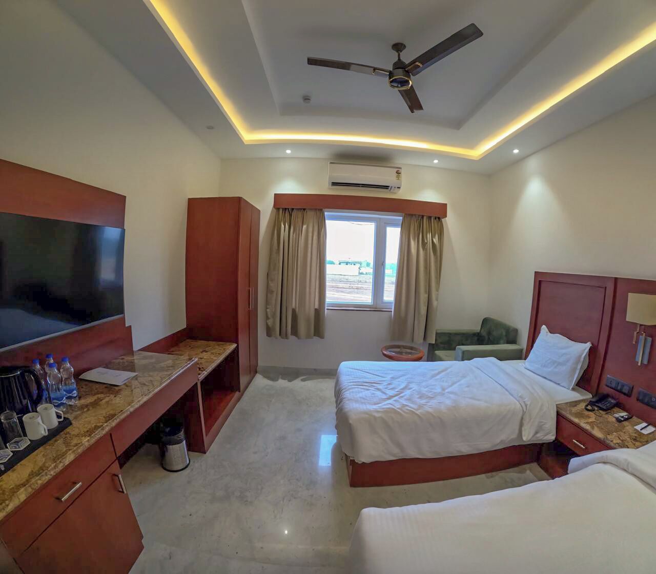 Una habitación de hotel con una cama, televisión, escritorio con computadora y ventilador de techo.