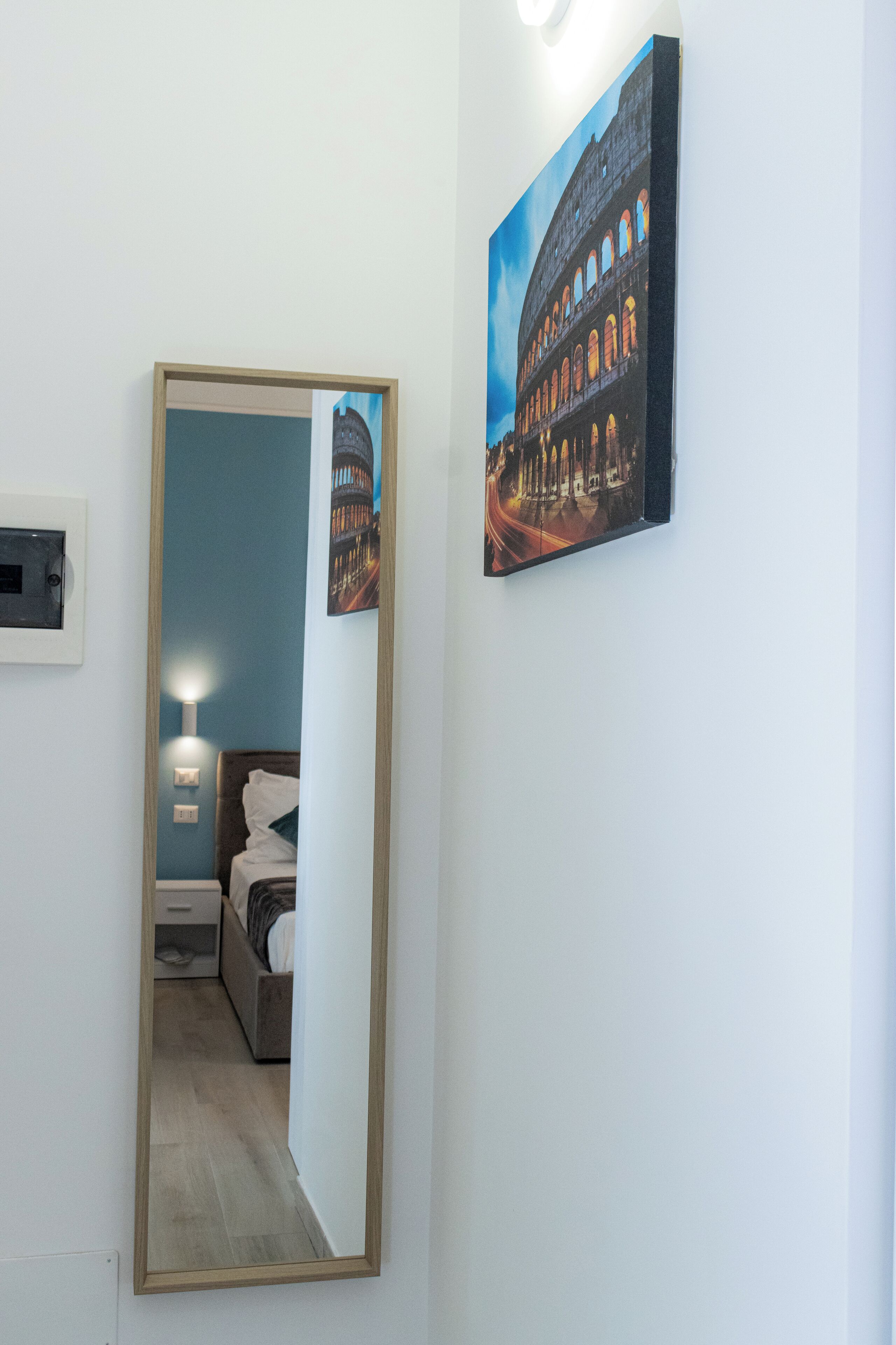 A hotel room with a bed, a nightstand, and a mirror reflecting the room. There is a painting of a building on the wall.