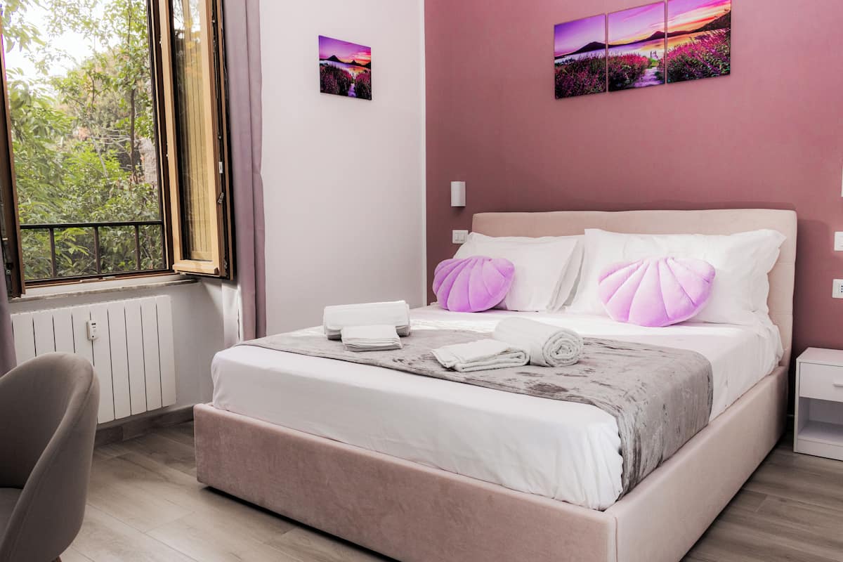 A neatly made bed with pillows, a bedside table, and a chair in a room with a window and artwork on the wall.