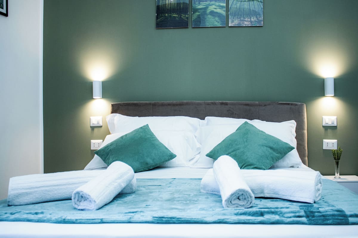 A neatly made bed with white linens and green pillows, two wall-mounted lights, and a small vase with a plant on the nightstand.