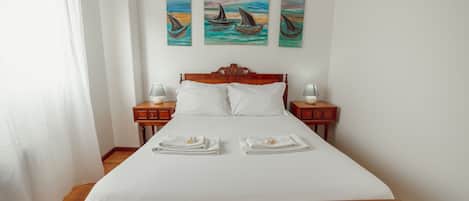Una cama bien hecha con ropa blanca, dos mesitas de noche con lámparas, y tres cuadros con barcos en la pared.