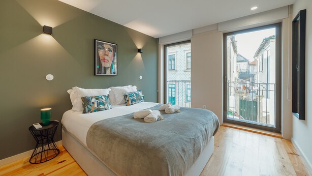 A modern bedroom with a large bed, a small side table, a wall-mounted light, and a window with a view of buildings. - Gaia Premium B Apartment by Vacationy (Vila Nova de Gaia)