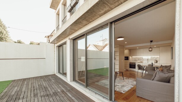 Un apartamento moderno con terraza de madera, puertas correderas de vidrio que dan a una cocina y una sala de estar, y un sofá gris.