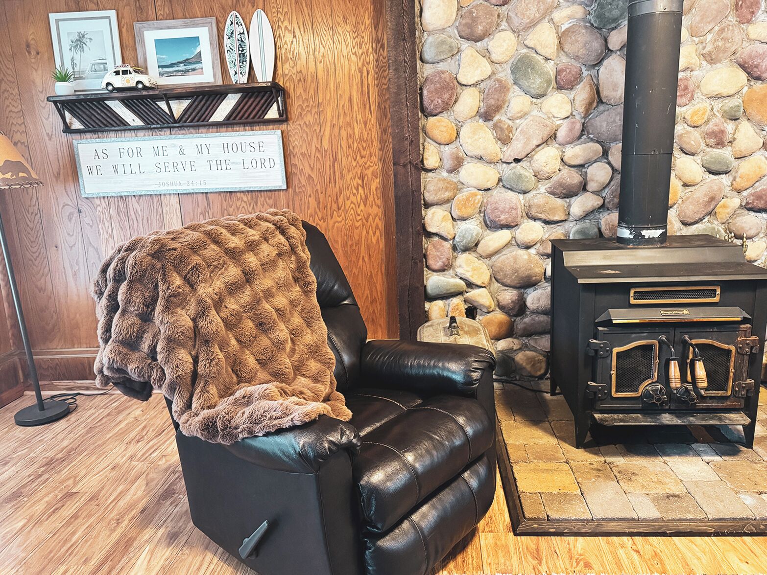 A cozy room with a leather chair covered with a blanket, a stone wall, a fireplace, and a shelf with framed pictures and a sign.