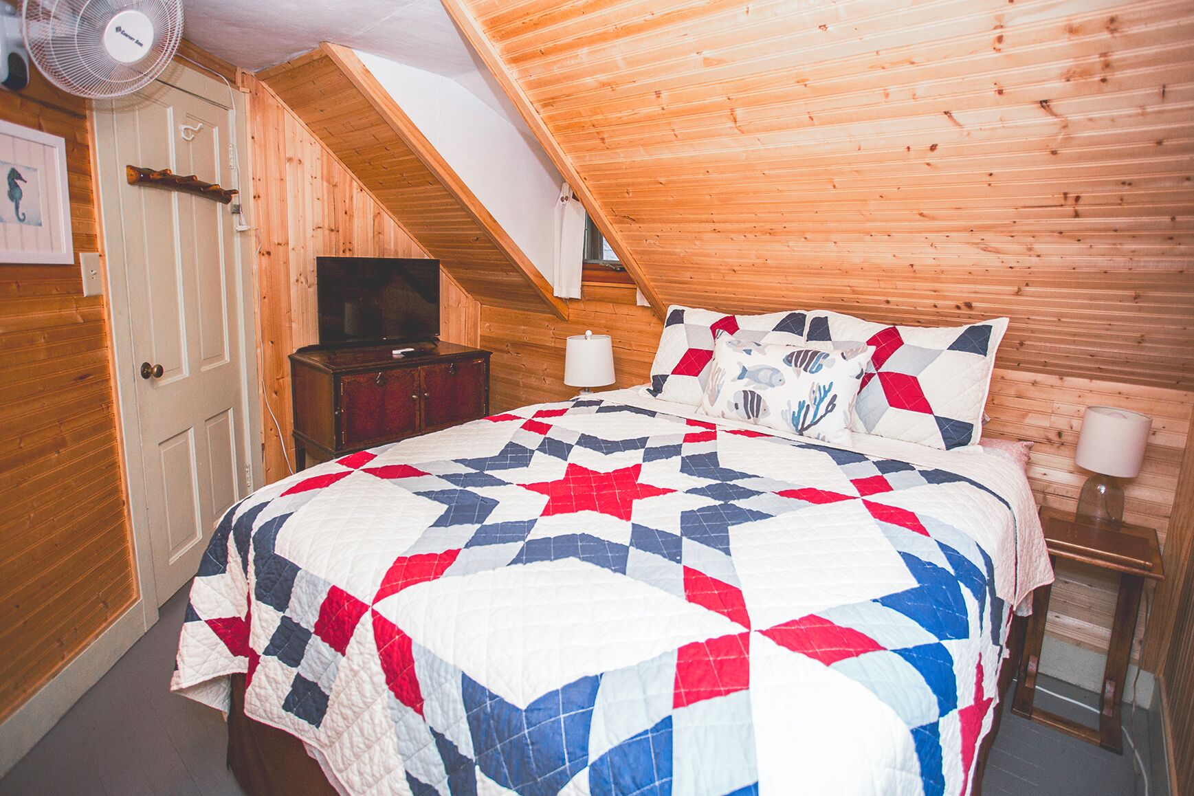 Une chambre mansardée chaleureuse avec un plafond en pente en bois, un lit recouvert d’une courtepointe à motifs d’étoiles, une télévision et une petite table avec une lampe.
