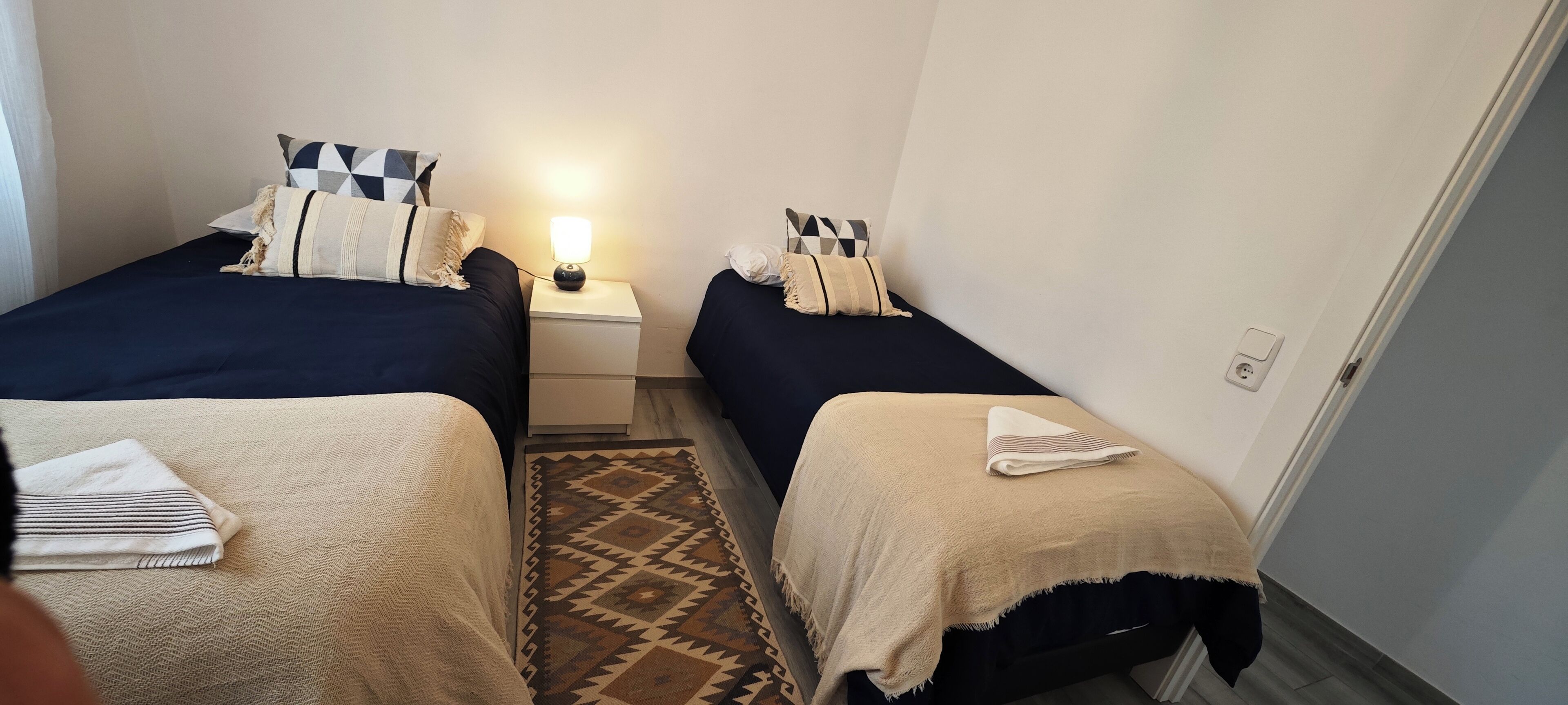 Una habitación de hotel pequeña con dos camas individuales, una mesita de noche con lámpara, un tapete estampado y una puerta a la derecha.