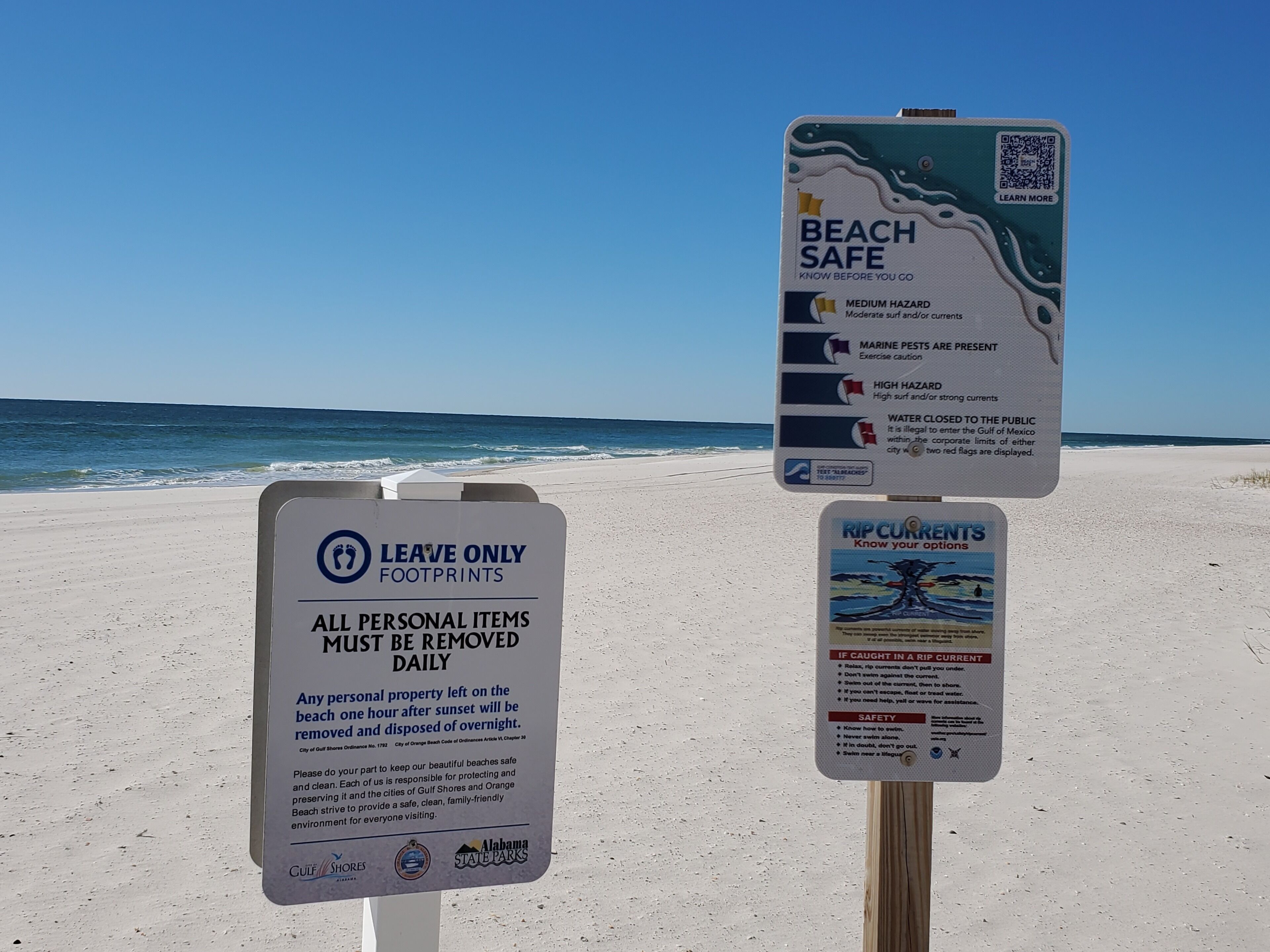 A beach with two informational signs. One sign warns of medium and high water hazards and rip currents. The other sign instructs to leave only footprints and remove personal items daily.