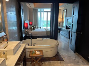 Une salle de bain moderne dotée d’une grande baignoire, d’une douche séparée et d’une chambre visible avec un lit et des tables de chevet.