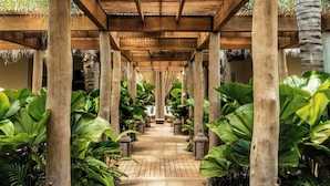 A wooden pathway flanked by large tropical plants and wooden pillars, leading to a covered seating area.