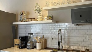 A modern kitchen with a white tiled backsplash, a stainless steel sink, and a coffee maker. There are shelves above the counter holding glassware and a vase with dried plants. - Taxqueño Charm  (Taxco de Alarcón)