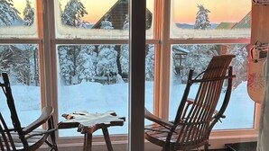 A room with large windows offering a view of a snowy landscape and a wooden cabin. There are two wooden chairs and a small table in the foreground. - A Perfect Mountain Getaway with Hot Tubs, and steps to Soaring Eagle ski lift! (Snowshoe)