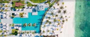 An aerial view of a resort with a large pool, beach area, and palm trees.