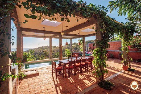Un espace extérieur abrité, aménagé pour les repas, avec une piscine, entouré de verdure et offrant une vue sur le paysage.