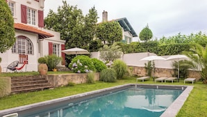Una piscina circondata da un giardino, scale che conducono a un edificio con persiane rosse e area relax all'aperto con ombrelloni.