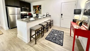 A modern kitchen with a central island, bar stools, stainless steel appliances, and a red side table with decorative items. - The Spot that Ever Calls - Walk to OM & Square! (Oxford)