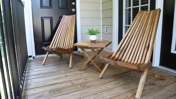 Two wooden Adirondack chairs with a small table on a wooden deck in front of a house with a black door and white siding.