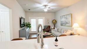 A modern kitchen with a white countertop, stainless steel sink, and a unique curved faucet. There is a dining area with a table and chairs, a sofa, and a lamp. A ceiling fan and a painting are visible on the walls.