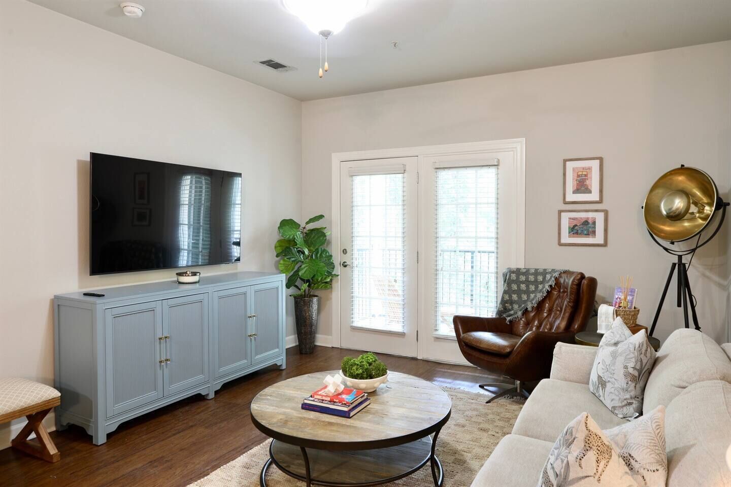 A living room with a flat-screen TV, a blue cabinet, a round coffee table with books, a brown leather chair, and a floor lamp.