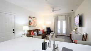Une chambre d’hôtel moderne avec un îlot de cuisine en marbre blanc, un évier intégré, un ventilateur de plafond, un canapé avec des coussins, une petite table ronde et un téléviseur fixé au mur.