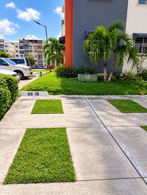 A parking area with marked spaces, a concrete pathway, and a building with a palm tree in the foreground. - Comodidad, seguridad y cercano a todo (Santo Domingo Este)