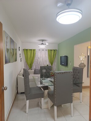 A dining area with a glass table, grey upholstered chairs, a white sofa, and a television on the wall. - Comodidad, seguridad y cercano a todo (Santo Domingo Este)