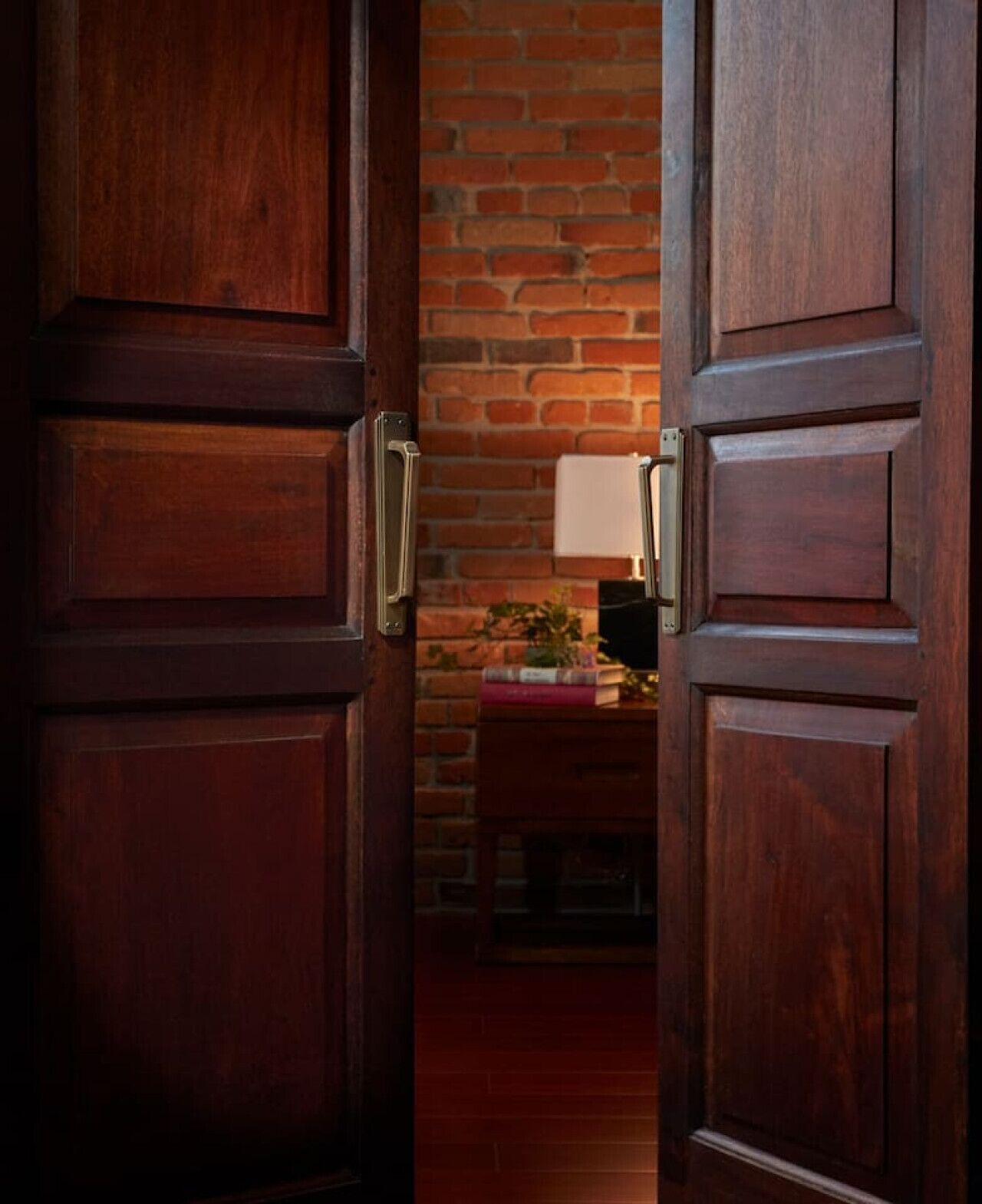 A wooden door with a metal handle, leading to a room with a brick wall and a small table with books and a lamp.