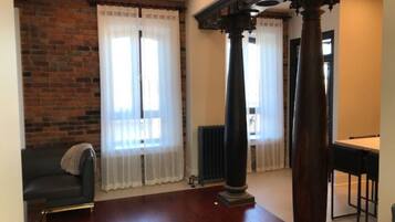 Een kamer met een bakstenen muur, twee grote ramen met witte gordijnen, een donker houten vloer, een zwarte leren bank en een zithoek met een tafel en stoelen.