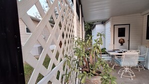 Ein überdachter Patio-Bereich mit weißem Gitterzaun, einem Topfpflanzen und Gartenmöbeln, darunter ein Tisch und Stühle.