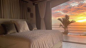 Un dormitorio con una cama grande, vistas al atardecer sobre el océano y una planta en maceta junto a la ventana.