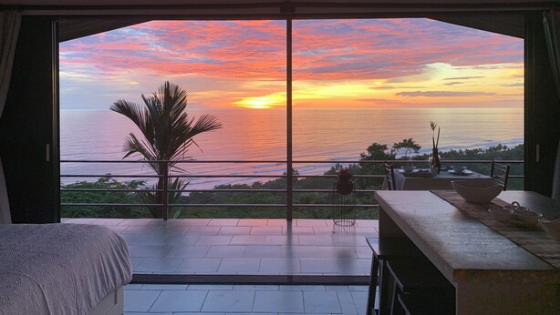 Una habitación con un ventanal que da a un atardecer sobre el océano, una cama y un comedor con mesa y sillas.