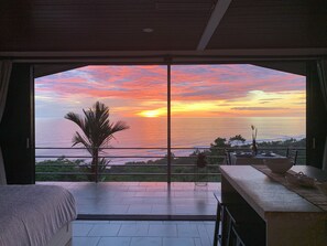 A room with a large window overlooking a sunset over the ocean, a bed, and a dining area with a table and chairs. - Villa Del Mar 3. 180 Ocean View, Gated community (Savegre de Aguirre)