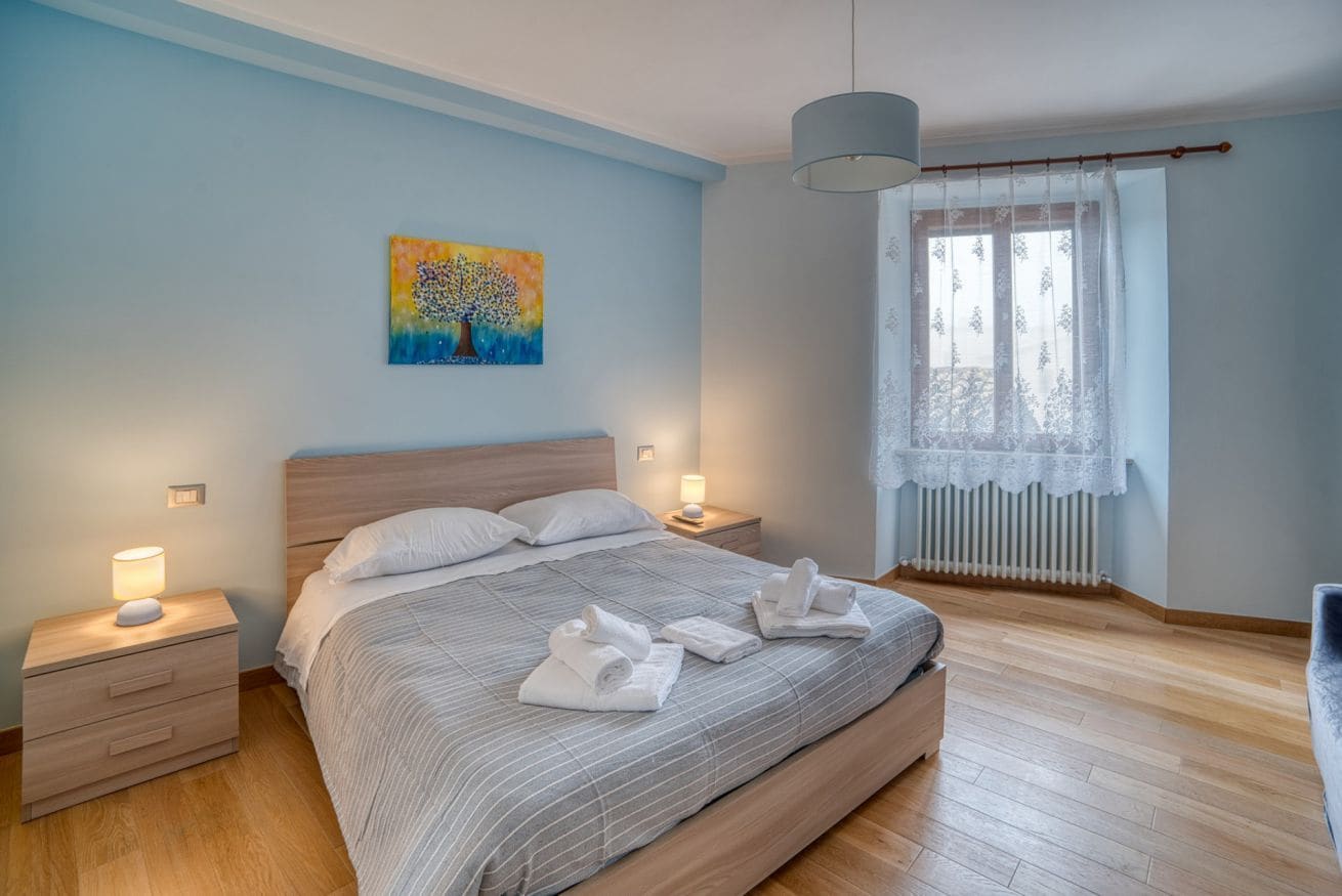 A bedroom with a bed, two bedside tables, a night lamp, a painting, a window with lace curtains, and a radiator.