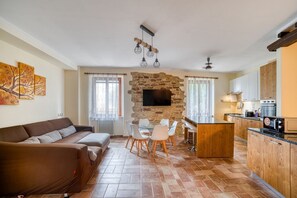 Una sala de estar con sofá, mesa de comedor, sillas, chimenea de piedra, televisor y área de cocina con armarios y electrodomésticos.