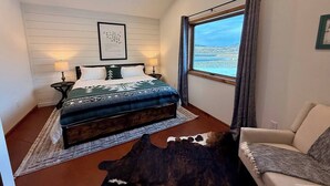A bedroom with a bed, a ceiling fan, a window with a view, a chair, and a rug. - Earhart Cabin at Ever Sky Ranch (Shell)