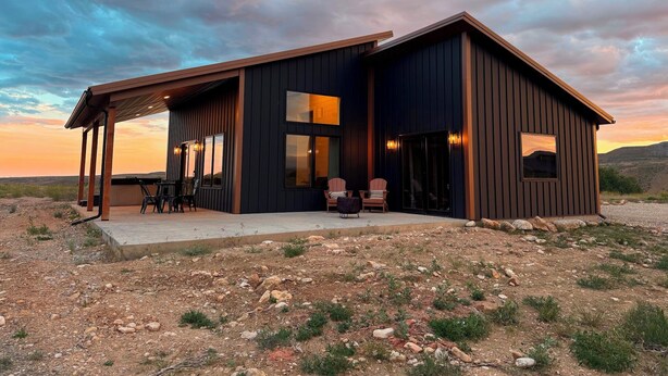 A modern house with a covered patio, large windows, and a view of the surrounding landscape. - Earhart Cabin at Ever Sky Ranch (Shell)